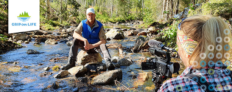 Inspelning av filmen Restaurering av vattendrag. En person sitter på en sten i ett vattendrag medan en fotograf ställer filmkameran. Foto: Tobias Eriksson