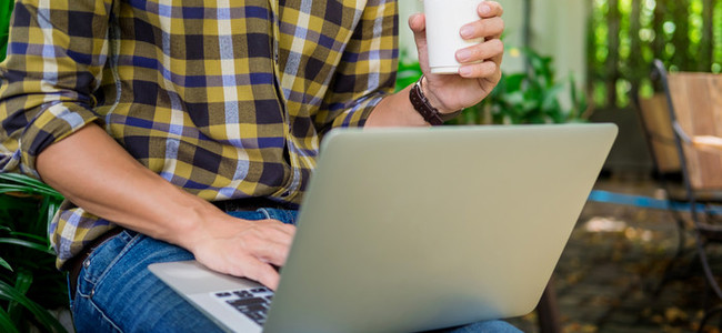 En man sitter utomhus med en laptop i knäet och en kaffemugg i handen. Foto: MostPhotos
