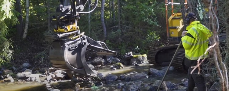 En grävmaskin arbetar med vattendragsrestaurering i ett skogsvattendrag. En arbetsledare i gul varseljacka står bredvid.  Foto: Länsstyrelsen Västerbotten
