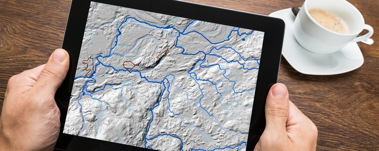 Närbild på en läsplatta. Någon håller plattan i båda händerna. På skärmen visas en vy från Skogsvattenkartan. Foto: Mostphotos, montage