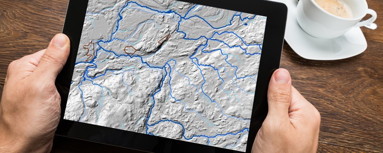 Närbild på en läsplatta. Någon håller plattan i båda händerna. På skärmen visas en vy från Skogsvattenkartan. Foto: Mostphotos, montage