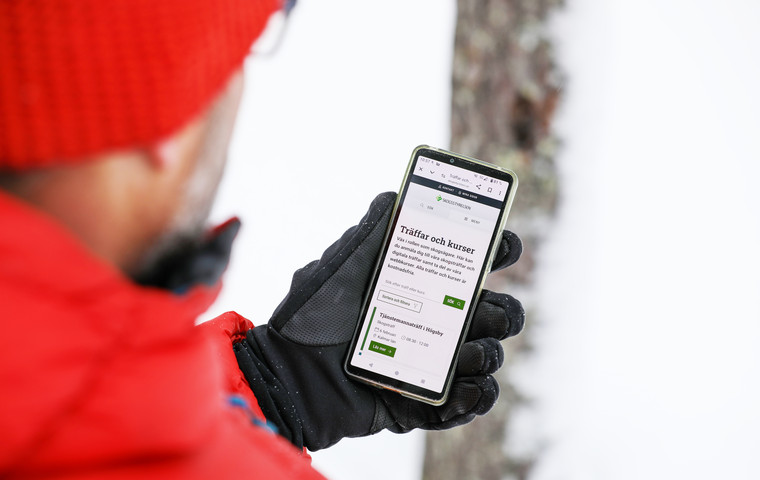 Närbild på en man i röd dunjacka och mössa som tittar på sin mobiltelefon. Fokus ligger på telefonen som mannen håller i sin hand. Foto: André Nyström