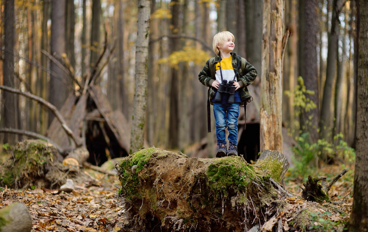 Liten kille med kikare på utflykt i skogen. I bakgrunden finns en koja. Foto: Maria Sbytova  / MostPhotos