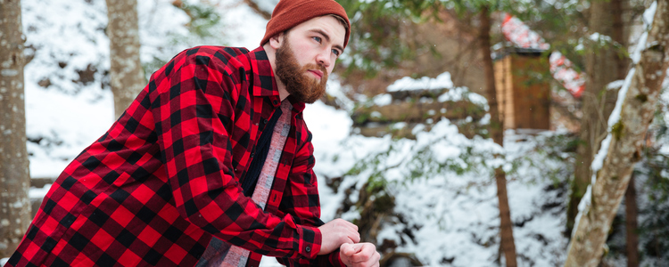 Man med skägg, rödrutig skjorta och mössa står och hänger på ett broräcke i en vintrig skog. . Foto: MostPhotos