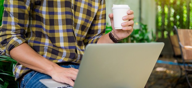Man som sitter framför en bärbar dator ute i skogen med en kopp kaffe i handen.