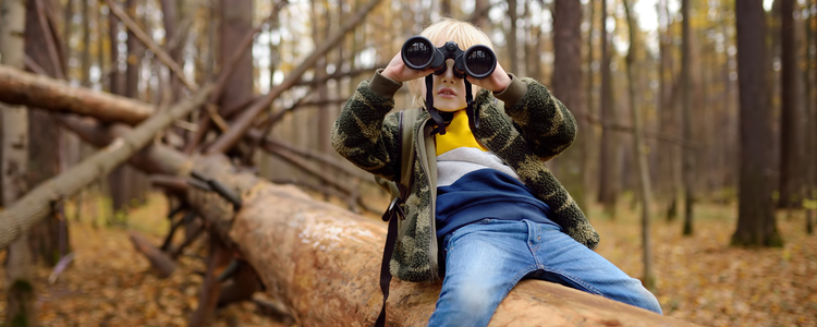 Liten kille med kikare på utflykt i skogen. Han sitter på en trädstam och kikar i sin kikare. Foto: Maria Sbytova  / MostPhotos