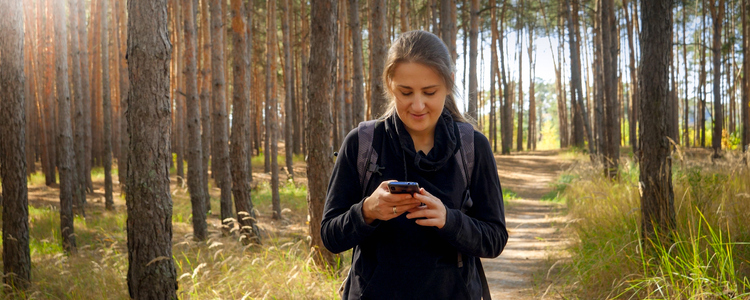 Ung kvinna som går i skogen, tittar ner på sin mobiltelefon. Foto: MostPhotos